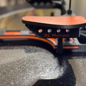 May include: Close-up of a black and orange RC Hangar component. The component has a textured orange surface and the text "RC Hangar" is visible. The design features geometric shapes and is set against a blurred background.