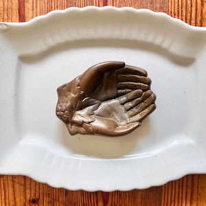 May include: A vintage bronze trinket dish shaped like two hands cupped together. The dish is on a white ceramic plate.