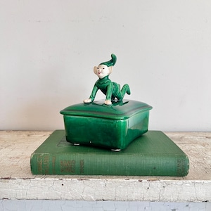 May include: A green ceramic trinket box with a lid, topped with a figurine of a crawling elf. The box is rectangular with rounded corners and sits on a green book. The elf is wearing a green outfit with a pointed hat and has a smiling face.
