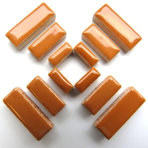 May include: A collection of rectangular, glossy, orange-brown tiles arranged on a white surface. The tiles have a smooth, reflective surface and a light-colored, textured backing. The arrangement suggests a mosaic or decorative tile design.