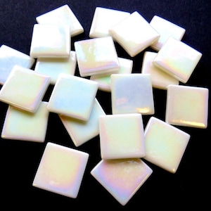 May include: A collection of iridescent square tiles. The tiles have a pearlescent finish that reflects a spectrum of colours, including yellow, pink, and blue. The tiles are arranged in a scattered pattern against a dark background.