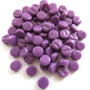 May include: A pile of glossy, purple glass beads. Some beads are smooth and round, while others have a textured, ridged design. The beads are scattered on a white surface, creating a visually appealing arrangement. These beads are suitable for various craft projects.