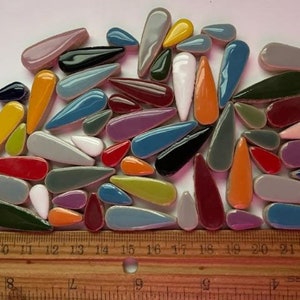 May include: A collection of colourful ceramic teardrop-shaped tiles in various shades of blue, green, red, yellow, orange, purple, and grey. The tiles are arranged in a random pattern and measure approximately 2.5 cm in length.