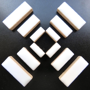 May include: Arrangement of white rectangular ceramic tiles with tan edges, set against a black background. The tiles are arranged in a symmetrical pattern, with larger tiles surrounding smaller ones. The overall composition is clean and minimalist.