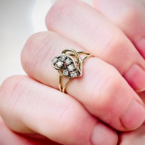 May include: A gold ring with a cluster of small, clear gemstones in a floral or leaf design. The ring has a delicate, openwork band. The ring is shown on a finger.
