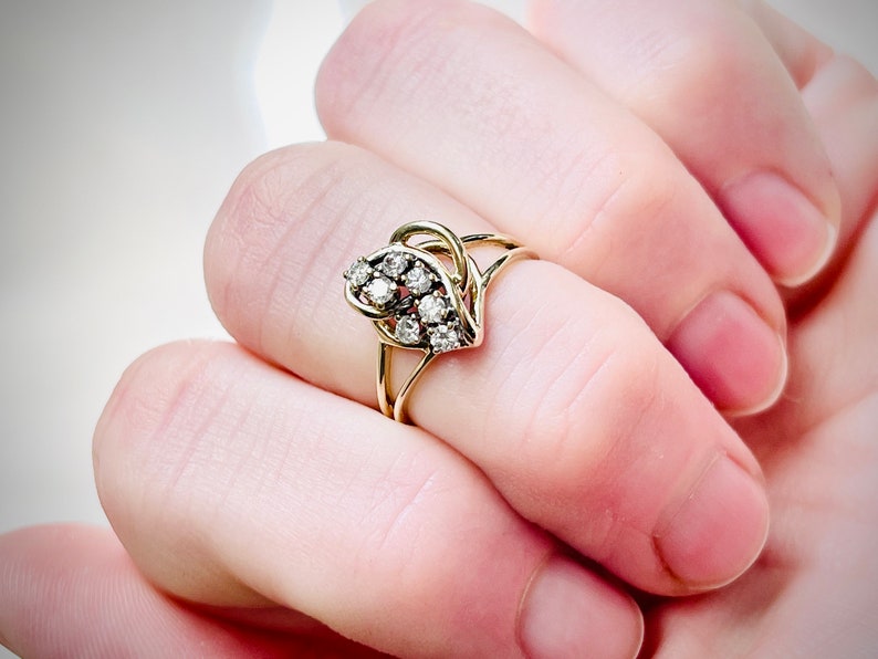 May include: A gold ring with a cluster of small, clear gemstones in a floral or leaf design. The ring has a delicate, openwork band. The ring is shown on a finger.