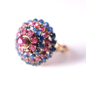 May include: A gold ring with a round, multi-colored gemstone setting. The gemstone is made up of small, red and blue stones.