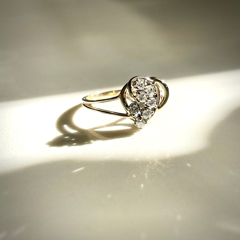 May include: A gold ring featuring a cluster of small, round, clear gemstones. The ring has a split shank design and a decorative setting for the gemstones. The ring is set against a white background with a shadow.