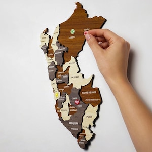 May include: A wooden wall map of Peru featuring distinct regions in varying shades of brown and cream. The map includes engraved text labels for provinces and cities, with small colorful circular markers placed on specific locations like Loreto and Cusco.