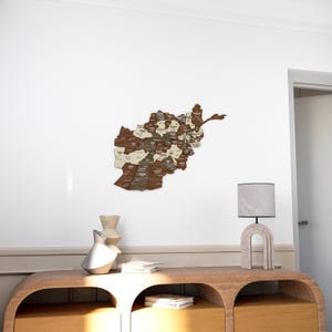 K&ouml;nnte beinhalten: Eine Holzkarte von Afghanistan mit den Provinzen in wei&szlig;er Schrift beschriftet. Die Karte ist an einer wei&szlig;en Wand &uuml;ber einem Holztisch mit zwei Schubladen angebracht. Eine Lampe mit einem grauen Schirm steht auf dem Tisch.