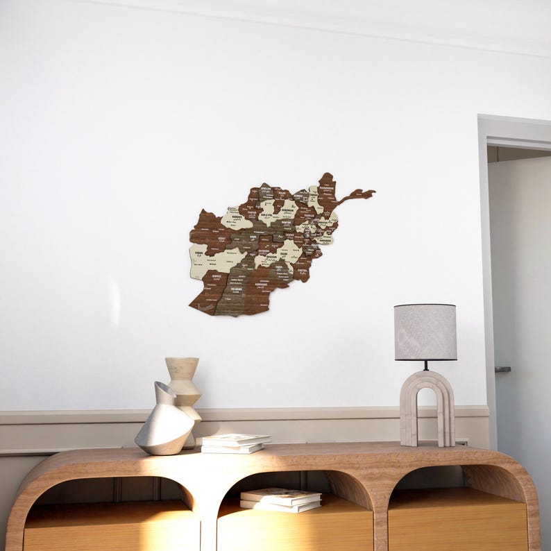 K&ouml;nnte beinhalten: Eine Holzkarte von Afghanistan mit den Provinzen in wei&szlig;er Schrift beschriftet. Die Karte ist an einer wei&szlig;en Wand &uuml;ber einem Holztisch mit zwei Schubladen angebracht. Eine Lampe mit einem grauen Schirm steht auf dem Tisch.
