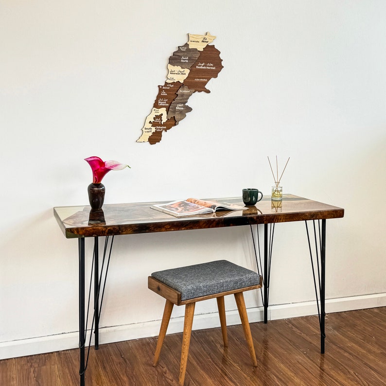 May include: A wooden map of Lebanon with the names of each region in Arabic and English. The map is hanging on a white wall above a dark brown wooden desk with black metal legs. A small gray stool sits in front of the desk.