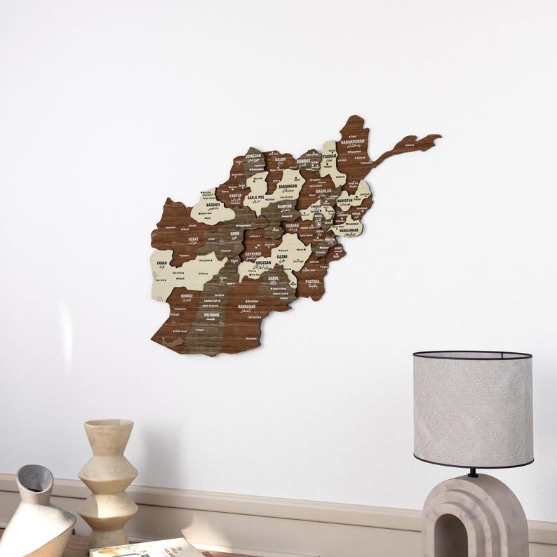 K&ouml;nnte beinhalten: Eine Holzkarte von Afghanistan mit den Namen der einzelnen Provinzen beschriftet. Die Karte ist an einer wei&szlig;en Wand mit einem beigefarbenen Sockel befestigt. Im Vordergrund steht eine Lampe mit einem grauen Stoffschirm auf einem beigefarbenen Tisch.