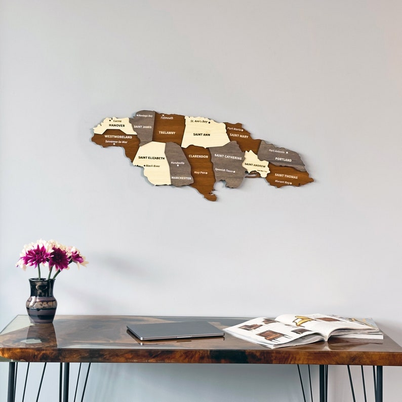 May include: A wooden map of Jamaica with the names of each parish labeled in white text. The map is mounted on a white wall above a wooden desk with a laptop and a magazine on top.