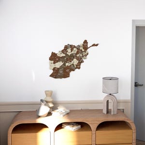 K&ouml;nnte beinhalten: Eine Holzkarte von Afghanistan mit den Provinzen in wei&szlig;er Schrift beschriftet. Die Karte ist an einer wei&szlig;en Wand &uuml;ber einer Kommode aus Holz mit zwei Schubladen angebracht. Eine Lampe mit einem wei&szlig;en Schirm steht auf der Kommode.