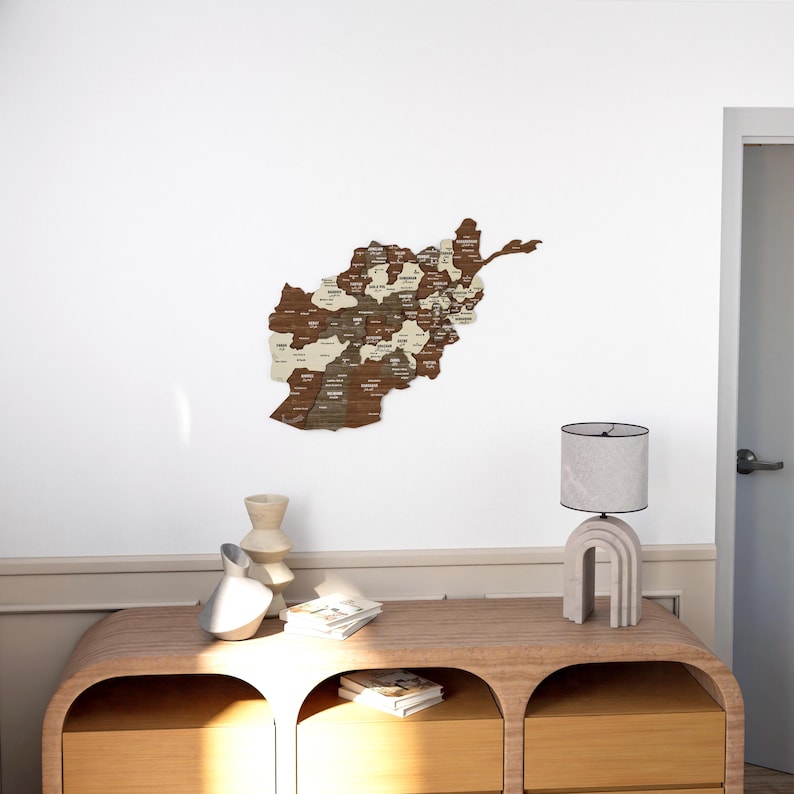 K&ouml;nnte beinhalten: Eine Holzkarte von Afghanistan mit den Provinzen in wei&szlig;er Schrift beschriftet. Die Karte ist an einer wei&szlig;en Wand &uuml;ber einer Kommode aus Holz mit zwei Schubladen angebracht. Eine Lampe mit einem wei&szlig;en Schirm steht auf der Kommode.