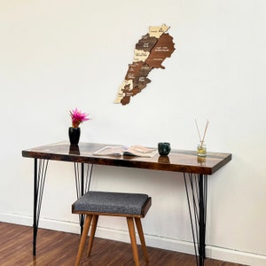 May include: A wooden map of Lebanon with the names of each region in Arabic. The map is hanging on a white wall above a dark brown desk with black metal legs. A small potted plant and a magazine are on the desk.