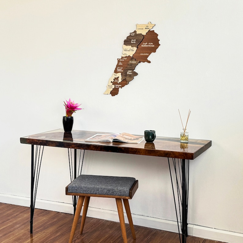 May include: A wooden map of Lebanon with the names of each region in Arabic. The map is hanging on a white wall above a dark brown desk with black metal legs. A small potted plant and a magazine are on the desk.