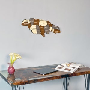 May include: A wooden map of Jamaica with the names of each parish labeled. The map is hanging on a light gray wall above a dark brown table with a laptop and a magazine on it. There are white flowers in a pink vase on the table.