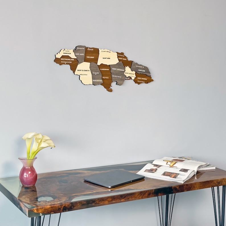 May include: A wooden map of Jamaica with the names of each parish labeled. The map is hanging on a light gray wall above a dark brown table with a laptop and a magazine on it. There are white flowers in a pink vase on the table.