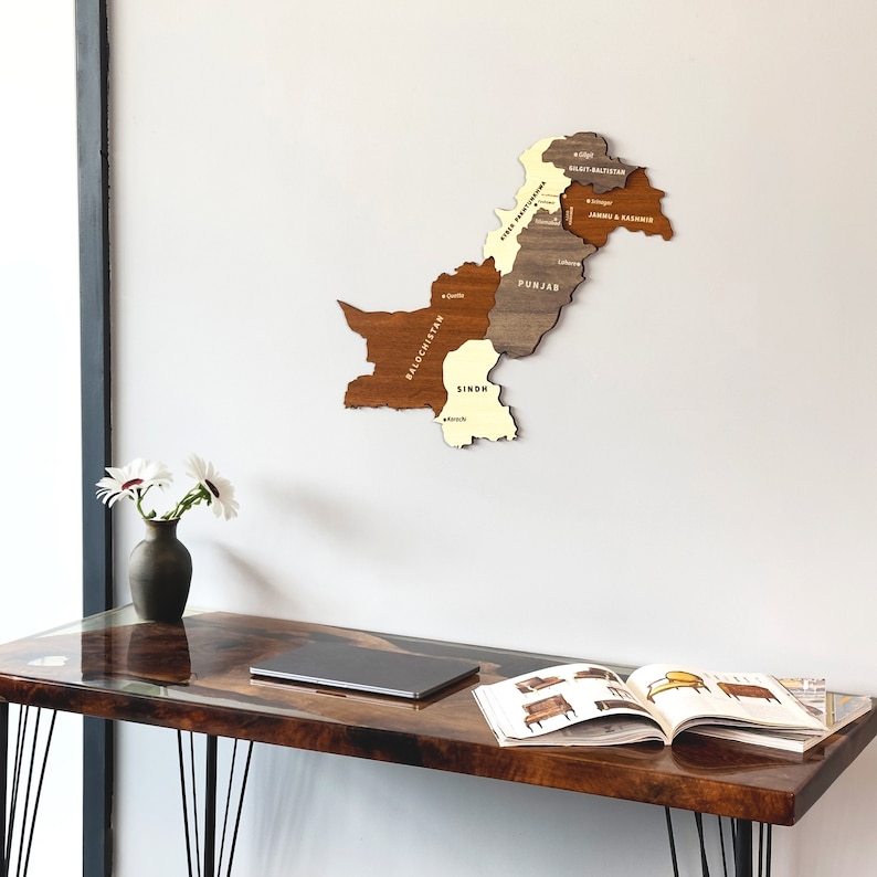 May include: A wooden map of Pakistan, showcasing the country's provinces in varying shades of brown and beige. The map is mounted on a white wall. The names of the provinces are printed on the map. A dark wooden desk with black hairpin legs is in the foreground.