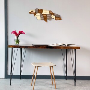 May include: A wooden map of Jamaica with the names of each parish labeled. The map is hanging on a white wall above a wooden desk with black hairpin legs. A laptop and a magazine are on the desk. A small beige stool is in front of the desk. A vase with pink calla lilies is on the desk.