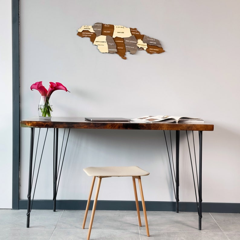 May include: A wooden map of Jamaica with the names of each parish labeled. The map is hanging on a white wall above a wooden desk with black hairpin legs. A laptop and a magazine are on the desk. A small beige stool is in front of the desk. A vase with pink calla lilies is on the desk.