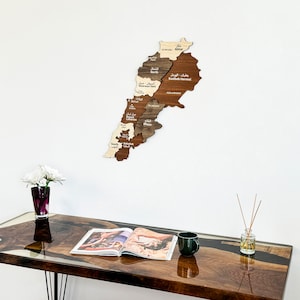 May include: A wooden map of Lebanon with the names of the different regions labeled in Arabic and English. The map is mounted on a white wall above a dark brown table with a magazine and a mug on it.