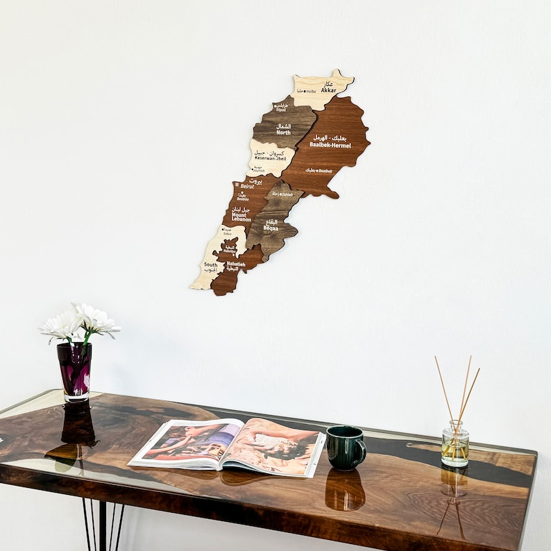 May include: A wooden map of Lebanon with the names of the different regions labeled in Arabic and English. The map is mounted on a white wall above a dark brown table with a magazine and a mug on it.