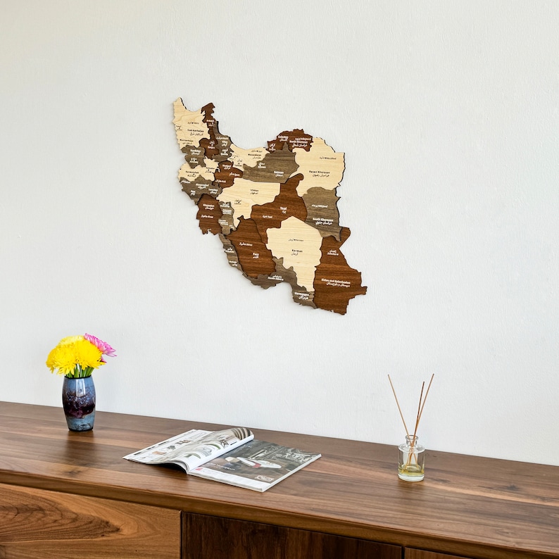 May include: A wooden map of Iran with the names of each province in Persian script. The map is mounted on a white wall and there is a vase of flowers and a magazine on a wooden table below it.