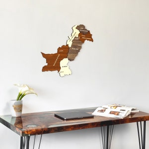 May include: A wooden map of Pakistan, showcasing its provinces in varying shades of brown and cream, mounted on a white wall. The map is above a desk with a glossy, wood-grain surface. A vase with a white flower and a laptop are also visible.
