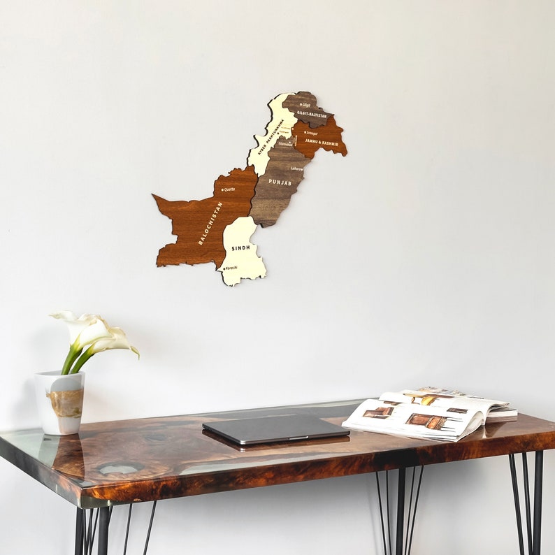 May include: A wooden map of Pakistan, showcasing its provinces in varying shades of brown and cream, mounted on a white wall. The map is above a desk with a glossy, wood-grain surface. A vase with a white flower and a laptop are also visible.