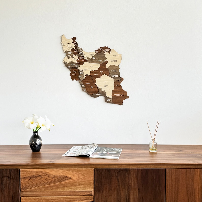 May include: A wooden map of Iran with the names of each province labeled. The map is mounted on a white wall above a wooden table with a magazine and a vase of white flowers.