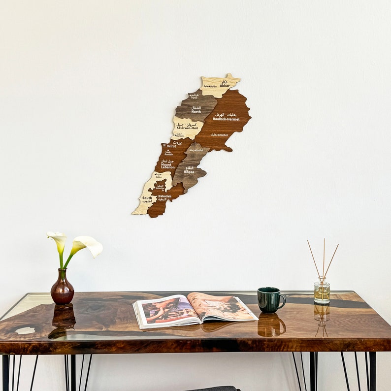 May include: A wooden map of Lebanon with the names of each region in Arabic and English. The map is mounted on a white wall above a dark brown table with a magazine and a green mug on it.