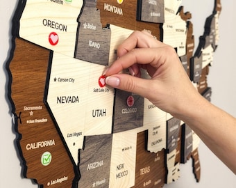 May include: A wooden wall map of the United States featuring laser-cut states in varying shades of light and dark wood. The map includes state names, city names, and small circular markers with heart, checkmark, and target icons placed on locations.