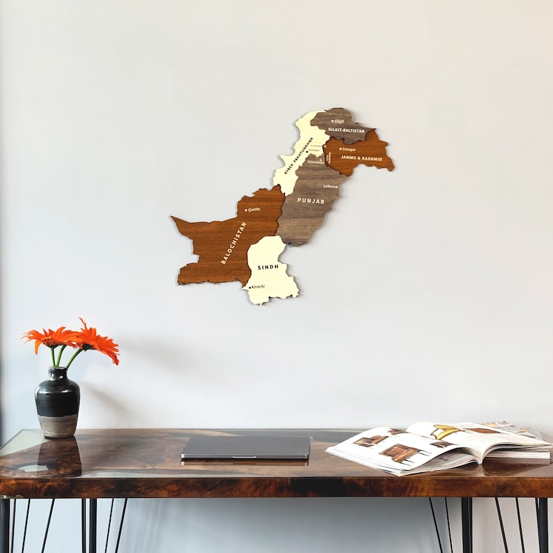 May include: A wooden map of Pakistan, showcasing the country's provinces in varying shades of brown and beige. The map is mounted on a white wall. A vase with orange flowers sits on a wooden desk in the foreground.