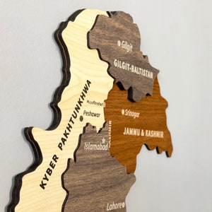 May include: A wooden map of Pakistan, featuring detailed cutouts of provinces in varying shades of brown and tan. Key cities like Islamabad, Lahore, and Peshawar are labeled. The map is mounted on a light gray surface.