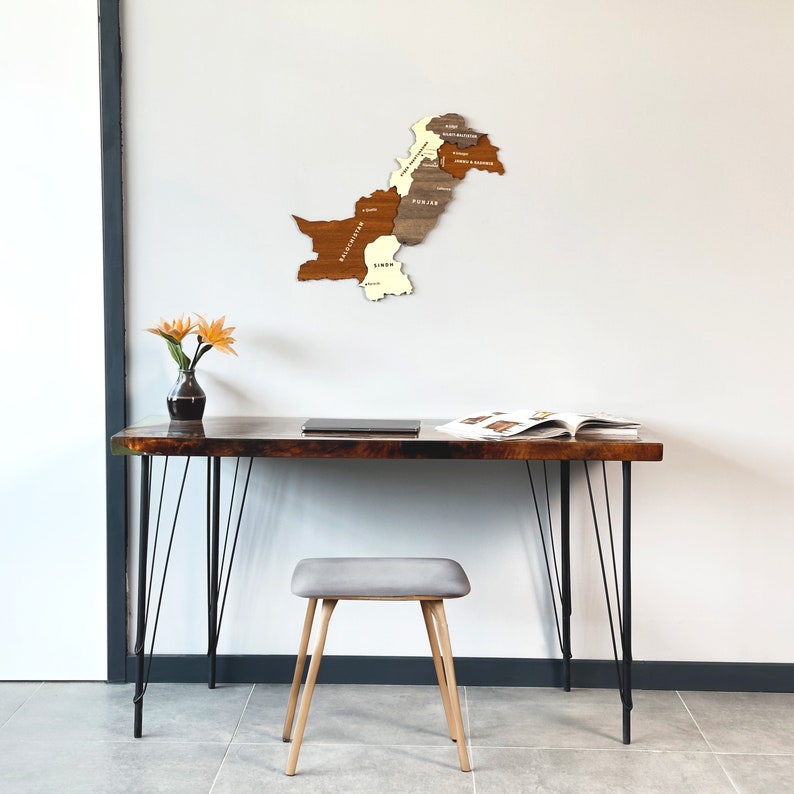 May include: A wooden map of Pakistan, segmented by province, hangs on a white wall. Below, a dark wood desk with black hairpin legs holds a laptop, open book, and a small vase with orange flowers. A gray stool with wooden legs sits in front.