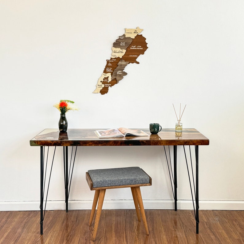 May include: A wooden map of Lebanon with the names of the different regions in Arabic and English. The map is hanging on a white wall above a brown desk with black metal legs. A gray stool sits in front of the desk.
