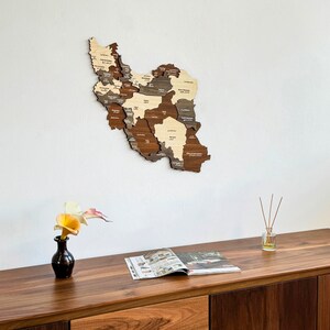 May include: A wooden map of Iran with the names of each province in both Farsi and English. The map is mounted on a white wall above a wooden table with a vase of flowers and a magazine.