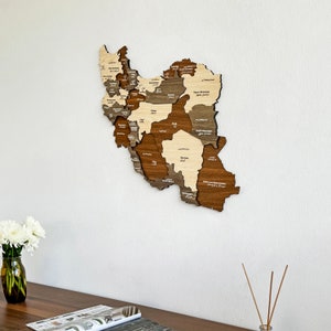May include: A wooden map of Iran with the names of each province in both Farsi and English. The map is mounted on a white wall and is surrounded by a white background.