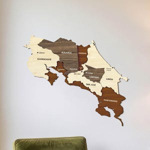 Może przedstawiać: Drewniana mapa Kostaryki na ścianę, przedstawiająca poszczególne prowincje w różnych odcieniach jasnego i ciemnego drewna. Mapa zawiera wygrawerowane nazwy regionów, takich jak Guanacaste, Alajuela, Heredia, Cartago, San José, Limón i Puntarenas.