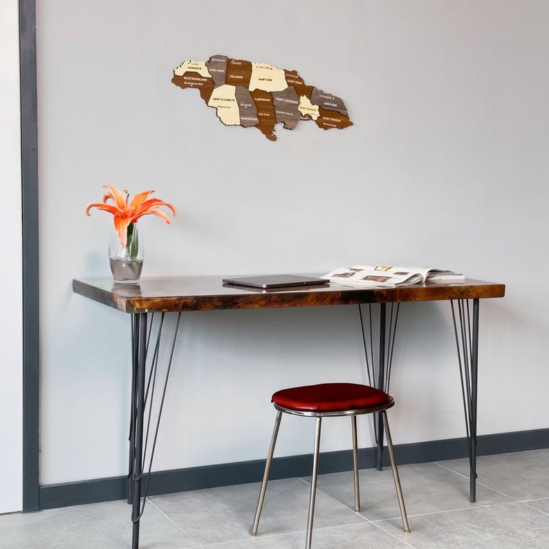 May include: A wooden map of Jamaica with the names of each parish, hanging on a white wall above a dark brown wooden desk with black metal hairpin legs. A red stool sits in front of the desk. A vase with orange flowers sits on the desk.