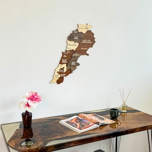 May include: A wooden map of Lebanon with the names of each region in Arabic. The map is mounted on a white wall above a dark brown wooden table with a magazine and a cup on it. A pink flower is in a vase on the table.