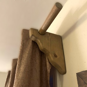 Puede incluir: Barra de cortina de madera con acabado marrón y soportes decorativos. La barra está montada en la pared y tiene una cortina marrón colgando de ella.