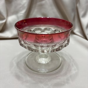 May include: A clear glass footed bowl with a ruby red rim. The bowl has a decorative pattern around the base and a circular design in the center. The bowl is likely vintage and suitable for serving desserts or displaying small items.