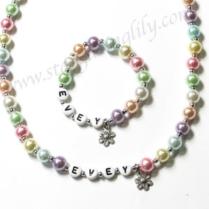 May include: A pastel-colored pearl necklace and bracelet set with a flower charm and the name "EVEY" spelled out in white letters.