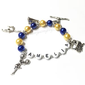 May include: A blue and gold beaded bracelet with a charm of a cheerleader holding a megaphone, a charm of a gymnast, and a charm of a cheerleader with the word "CHEER" on it. The bracelet also has white beads with the letters "AMELIA" on them.