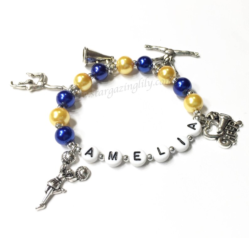 May include: A blue and gold beaded bracelet with a charm of a cheerleader holding a megaphone, a charm of a gymnast, and a charm of a cheerleader with the word "CHEER" on it. The bracelet also has white beads with the letters "AMELIA" on them.