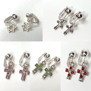 May include: A collection of clip-on earrings featuring various religious symbols. The earrings include silver-toned clip-on backs with dangling charms. The charms include a Star of David, crosses, and cross-shaped charms adorned with pink, green, and red gemstones.